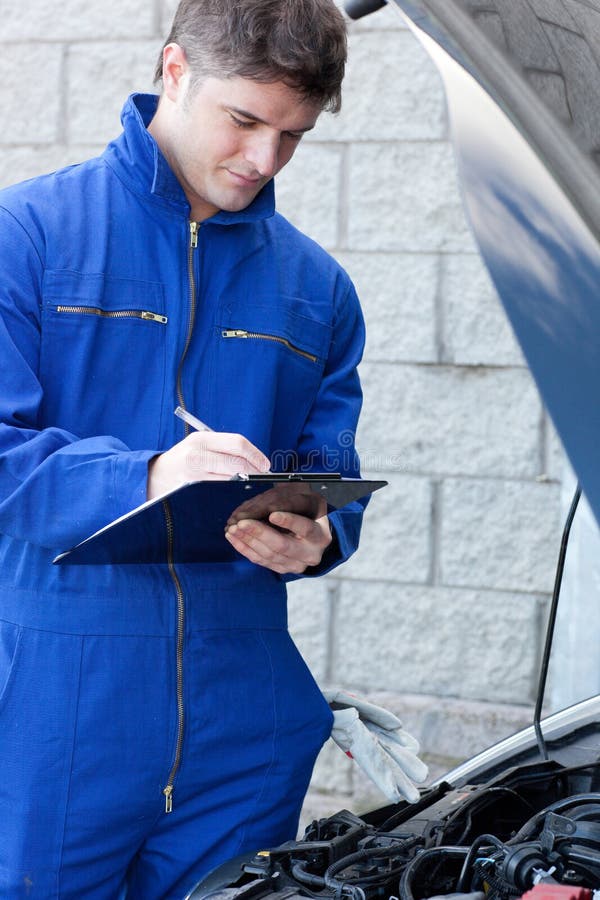 Handsome mechanic writing on a clipboard standing in front of a car at work. Handsome mechanic writing on a clipboard standing in front of a car at work