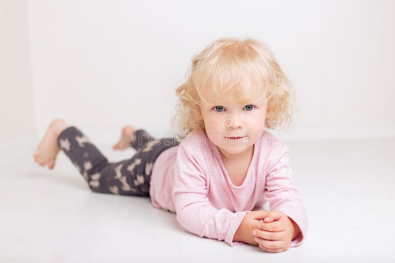 Het krullende meisje van de blondebaby in comfortabele huis vrijetijdskleding ligt op de vloer in een heldere witte ruimte glimla stock foto's Het krullende meisje van de blondebaby in comfortabele huis vrijetijdskleding ligt op de vloer in een heldere witte ruimte glimla stock foto's