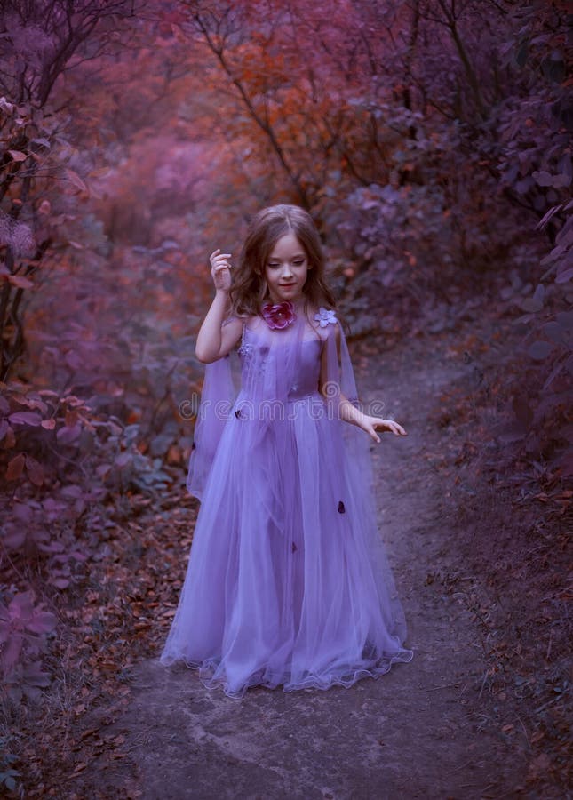 Het leuke meisje bevindt zich in het bos in een purpere lichte lange kleding met bloemen, een kleine prinses als in een droom, ga stock foto Het leuke meisje bevindt zich in het bos in een purpere lichte lange kleding met bloemen, een kleine prinses als in een droom, ga stock foto