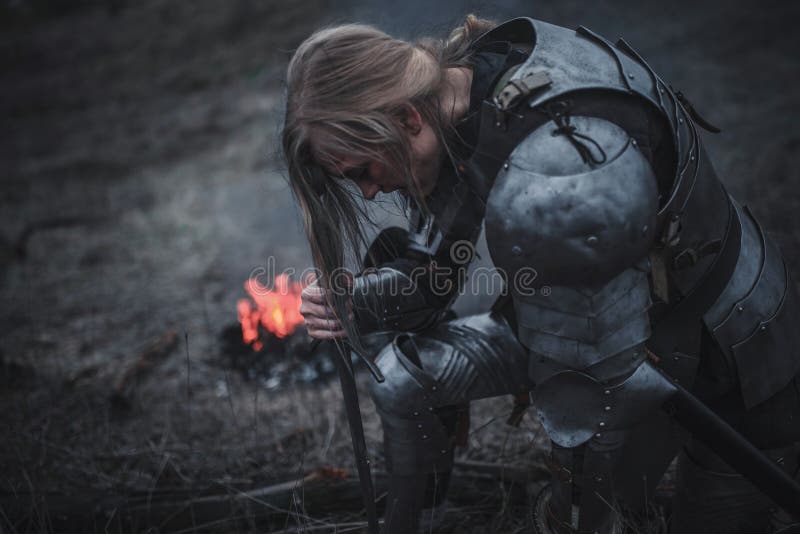 Het meisje in beeld van de Boog van Jeanne D ` knielt in pantser en met zwaard in haar handen tegen achtergrond van brand en rook royalty-vrije stock foto's Het meisje in beeld van de Boog van Jeanne D ` knielt in pantser en met zwaard in haar handen tegen achtergrond van brand en rook royalty-vrije stock foto's