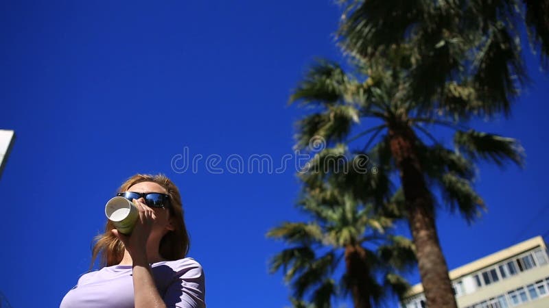 Het meisje drinkt een drank op de straat van een beschikbare kop tegen de blauwe hemel en de palmen Knippend inbegrepen weg