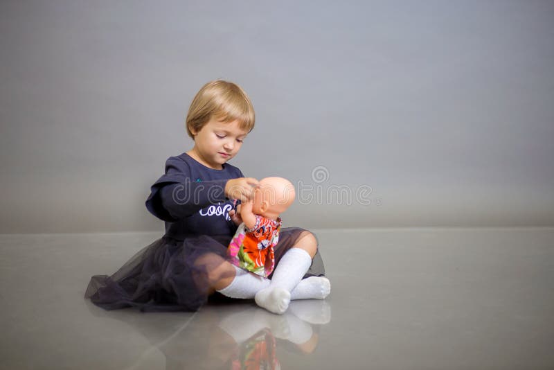 Het meisje in een grijze kleding zit op een grijze achtergrond en speelt met een poppenbaby - pop stock afbeeldingen Het meisje in een grijze kleding zit op een grijze achtergrond en speelt met een poppenbaby - pop stock afbeeldingen