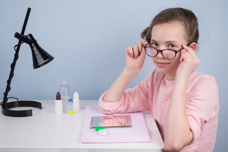 Het meisje stijgt haar glazen op, zittend bij de lijst om de lens te dragen voor het verbeteren van het gezicht stock afbeeldingen Het meisje stijgt haar glazen op, zittend bij de lijst om de lens te dragen voor het verbeteren van het gezicht stock afbeeldingen