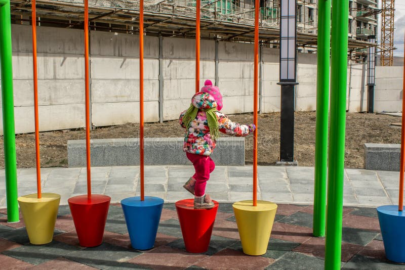 Het meisje, warm gekleed, in een hoed en jasjespelen op de speelplaats met dia's en schommeling in de binnenplaats van residentia stock foto Het meisje, warm gekleed, in een hoed en jasjespelen op de speelplaats met dia's en schommeling in de binnenplaats van residentia stock foto
