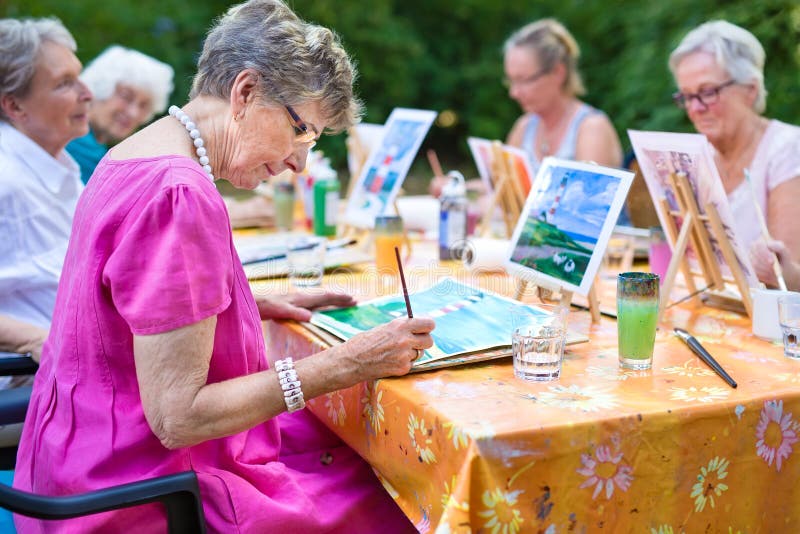 Het modieuze hogere dame schilderen in kunstklasse met vrienden van haar zorghuis voor oud kopiërend het schilderen met waterkleu
