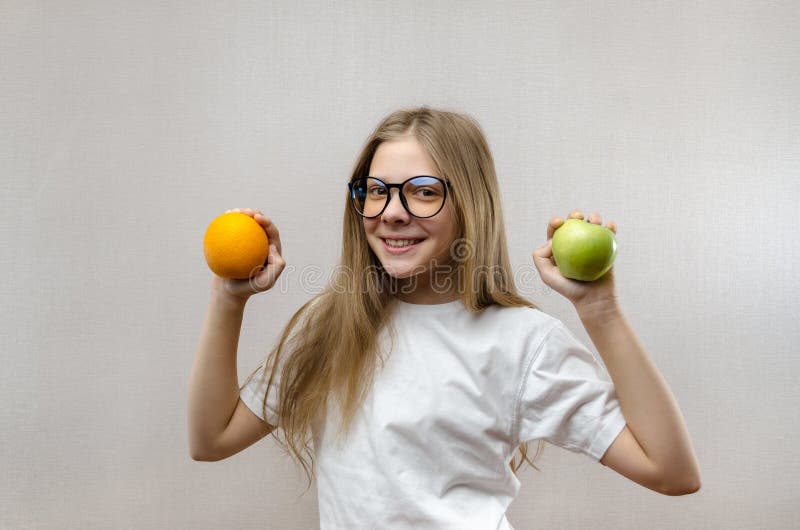 Het mooie blondemeisje in een witte T-shirt glimlacht en houdt een appel en een sinaasappel in haar handen Gezonde voeding voor royalty-vrije stock afbeelding Het mooie blondemeisje in een witte T-shirt glimlacht en houdt een appel en een sinaasappel in haar handen Gezonde voeding voor royalty-vrije stock afbeelding