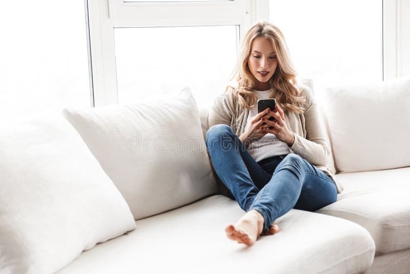 Het mooie blondevrouw stellen die thuis binnen gebruikend mobiele telefoon zitten stock fotografie Het mooie blondevrouw stellen die thuis binnen gebruikend mobiele telefoon zitten stock fotografie