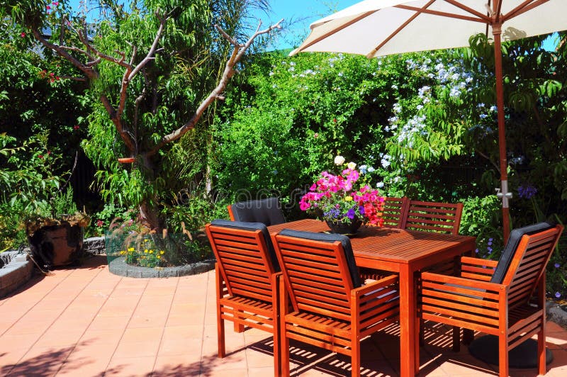 Beautiful and lush summertime Mediterranean style courtyard garden with wooden table and chairs and white market umbrella. Beautiful and lush summertime Mediterranean style courtyard garden with wooden table and chairs and white market umbrella.