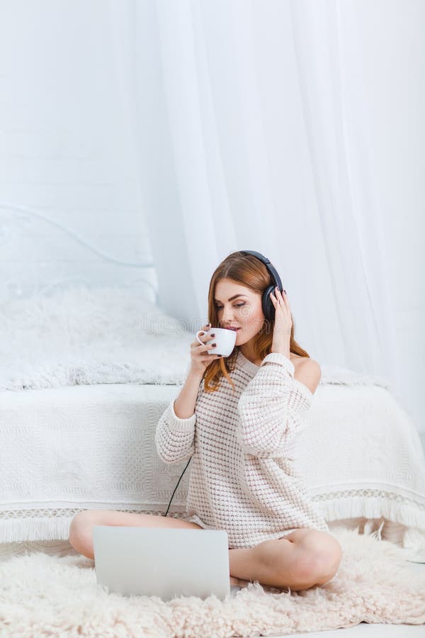 Het mooie meisje in een warme sweater, zit op de vloer dichtbij laptop met hoofdtelefoons, en drinkt thee binnen royalty-vrije stock afbeelding Het mooie meisje in een warme sweater, zit op de vloer dichtbij laptop met hoofdtelefoons, en drinkt thee binnen royalty-vrije stock afbeelding