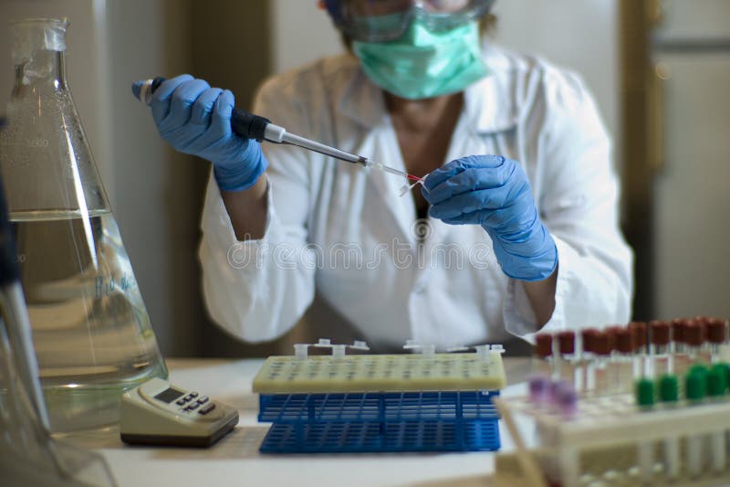 Is a scientific woman pipetting cells cultured in serum in an Eppendorf tube. The scientist wears a lab coat, gloves, safety goggles and mask. It is surrounded by laboratory equipment: tubes, racks, timer, pipetes. Is a scientific woman pipetting cells cultured in serum in an Eppendorf tube. The scientist wears a lab coat, gloves, safety goggles and mask. It is surrounded by laboratory equipment: tubes, racks, timer, pipetes