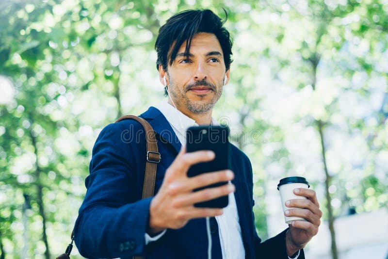 Het portret van modieuze jonge zakenman die smartphone voor het listining van muziek gebruiken terwijl het lopen in stadspark en royalty-vrije stock foto Het portret van modieuze jonge zakenman die smartphone voor het listining van muziek gebruiken terwijl het lopen in stadspark en royalty-vrije stock foto
