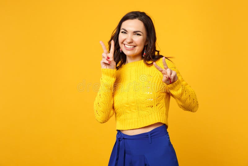 Het portret van vrolijke jonge vrouw in sweater, blauwe broeken die camera die overwinningsteken tonen isoleerde op geeloranje ki stock afbeelding Het portret van vrolijke jonge vrouw in sweater, blauwe broeken die camera die overwinningsteken tonen isoleerde op geeloranje ki stock afbeelding