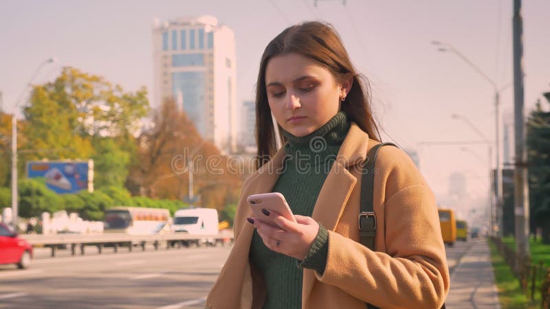 Het schitterende Kaukasische wijfje scrolt haar telefoon terwijl status dichtbij weg met verkeer, geconcentreerde stedelijke stat