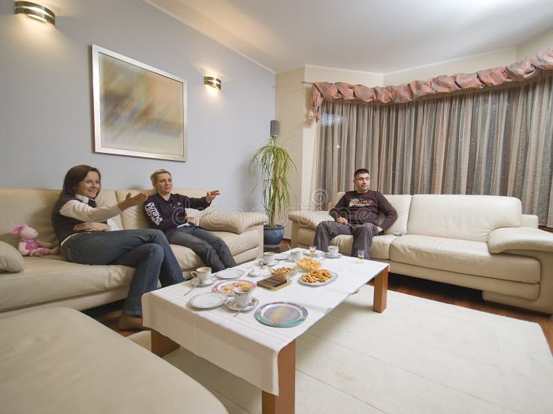 Three adults sitting on a sofa in the modern living room, talking. The table with some party snacks. All of them watching TV, making gestures. Three adults sitting on a sofa in the modern living room, talking. The table with some party snacks. All of them watching TV, making gestures.