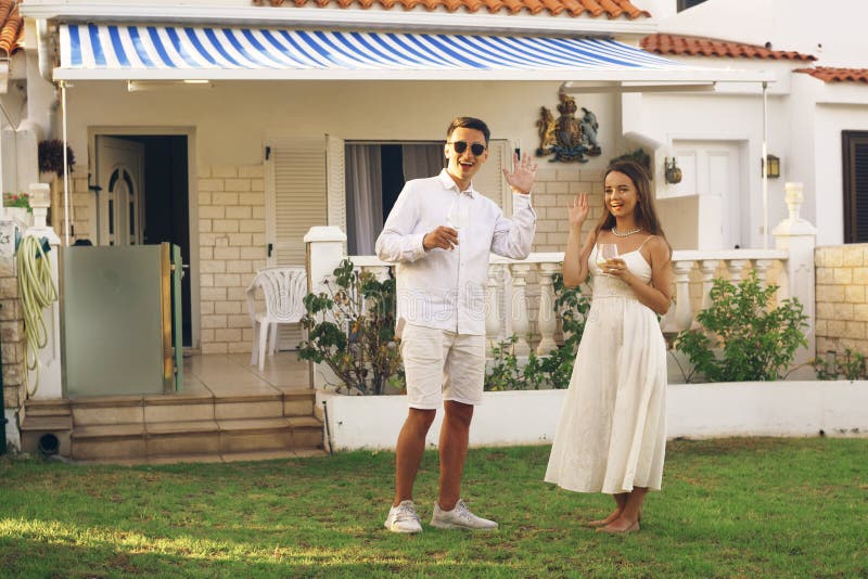 The couple just moved to a new house on the ocean, the couple stands on the lawn drinking wine and waving to the guests. The couple communicates with neighbors. Happy girl and guy drinks wine on vacation. Couple dressed in white. The couple just moved to a new house on the ocean, the couple stands on the lawn drinking wine and waving to the guests. The couple communicates with neighbors. Happy girl and guy drinks wine on vacation. Couple dressed in white