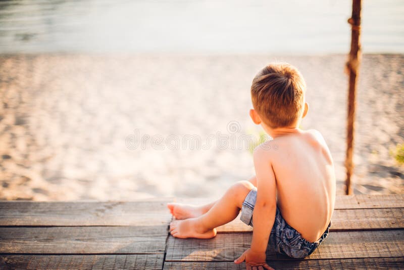 Het thema is een kind en de zomerstrandvakantie Een kleine jongen zit met zijn rug op een houten pijler en bewondert de mening va stock afbeelding Het thema is een kind en de zomerstrandvakantie Een kleine jongen zit met zijn rug op een houten pijler en bewondert de mening va stock afbeelding
