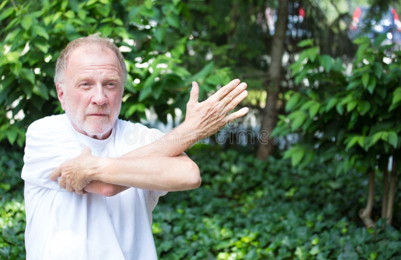 Closeup portrait, senior mature man in white shirt, stretching arms, isolated green trees background. Warming up. Closeup portrait, senior mature man in white shirt, stretching arms, isolated green trees background. Warming up