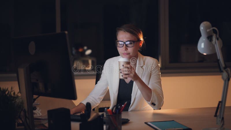 Het vermoeide blonde in glazen en kostuum werkt aan computer laat bij nacht en het drinken meeneem vermoeid koffiegevoel en