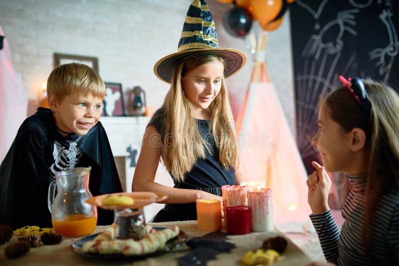 Profile view of pretty girl telling scary story to her little friends while celebrating Halloween at home, table set for holiday dinner on foreground. Profile view of pretty girl telling scary story to her little friends while celebrating Halloween at home, table set for holiday dinner on foreground