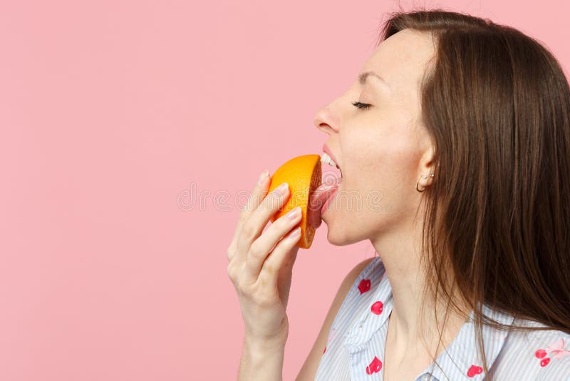 Het zijaanzicht van jonge vrouw in de zomerkleren die ogen houden sloot het likken van helft van vers oranje fruit die op roze wo stock foto's Het zijaanzicht van jonge vrouw in de zomerkleren die ogen houden sloot het likken van helft van vers oranje fruit die op roze wo stock foto's