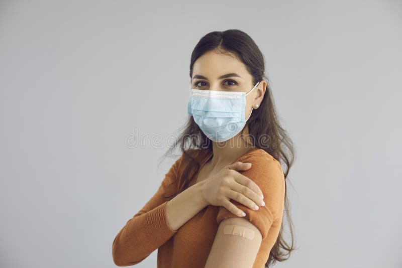 Happy beautiful young woman wearing medical face mask isolated on gray copy space background shows adhesive plaster on arm after getting safe Covid 19 vaccine. Coronavirus vaccination campaign concept. Happy beautiful young woman wearing medical face mask isolated on gray copy space background shows adhesive plaster on arm after getting safe Covid 19 vaccine. Coronavirus vaccination campaign concept