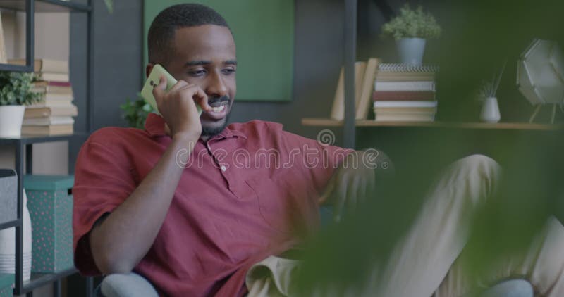 Heureux afro-américain parler sur téléphone portable assis dans un fauteuil dans l'appartement banque de vidéos