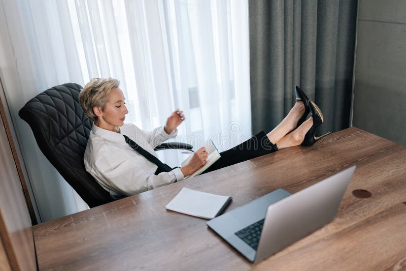 High-angle view of middle-aged female CEO putting feet on desk with laptop. Confident successful business woman thinking about career growth relaxing in cabinet. Concept relaxation at workplace. High-angle view of middle-aged female CEO putting feet on desk with laptop. Confident successful business woman thinking about career growth relaxing in cabinet. Concept relaxation at workplace