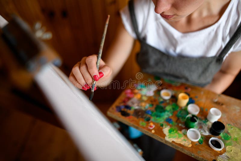 High angle view on woman artist with palette with and brush in her hand who paints on canvas. High angle view on woman artist with palette with and brush in her hand who paints on canvas