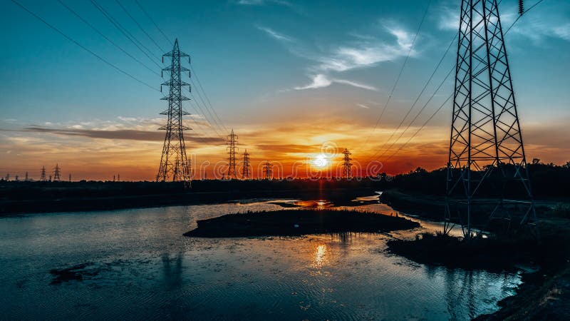 Silhouette effect of high voltage electric towers with electricity transmission power lines, located near riverside & sunset