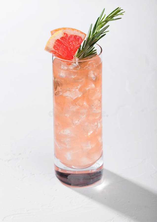 Highball glass of refreshing summer red grapefruit cocktail with ice cubes, fruit slice and rosemary on white background