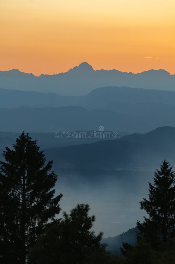 The highest peak of Julian Alps on orange sky with silhouette of spruce treetops in foreground stock photos The highest peak of Julian Alps on orange sky with silhouette of spruce treetops in foreground stock photos