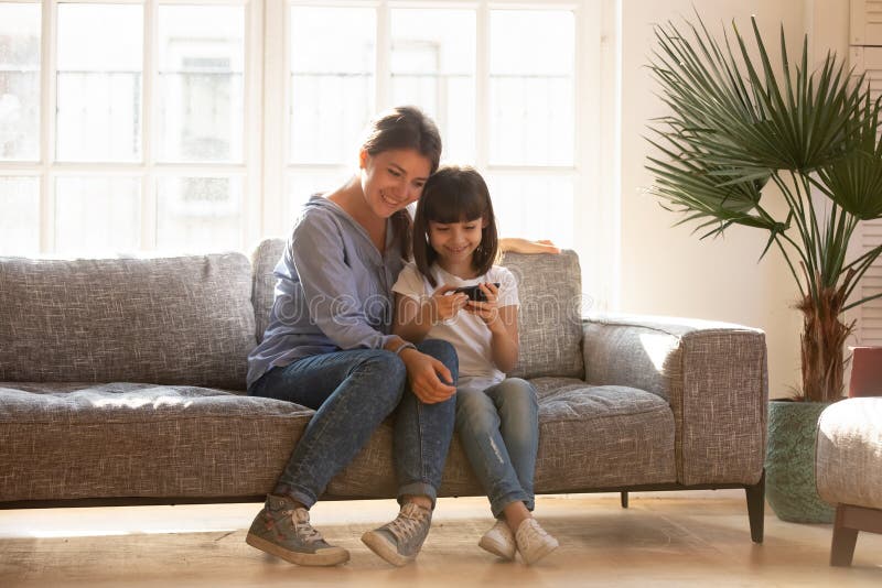 Hija feliz de la mamá y del niño que usa el smartphone que se sienta en el sofá foto de archivo Hija feliz de la mamá y del niño que usa el smartphone que se sienta en el sofá foto de archivo