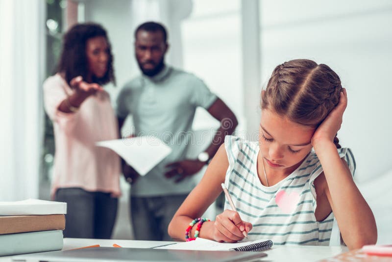 Hija triste que hace las tareas caseras mientras que padres que gritan en ella imagen de archivo Hija triste que hace las tareas caseras mientras que padres que gritan en ella imagen de archivo