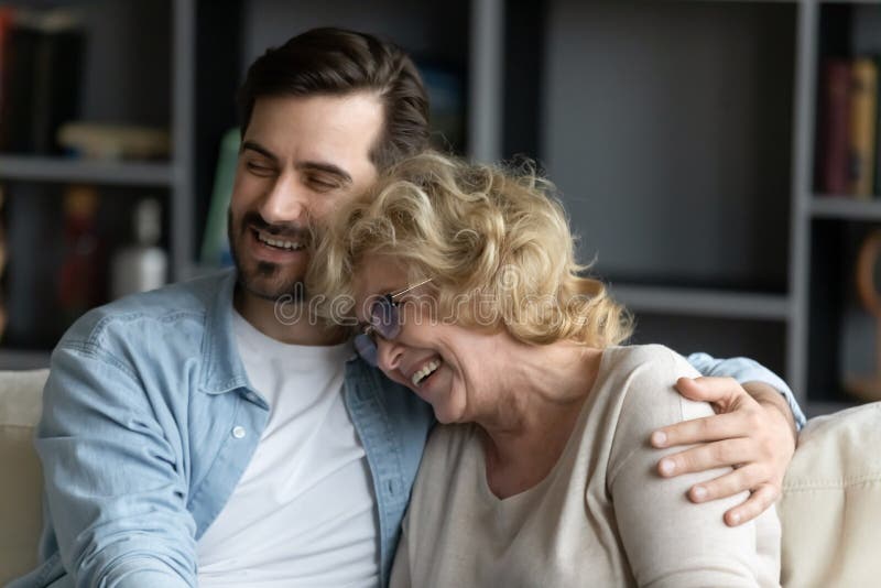 Close up of grown up child son hugs elderly mother family sit on couch enjoy talk funny conversation laughing, remember life, share positive news feels happy spend time together, family bonds concept. Close up of grown up child son hugs elderly mother family sit on couch enjoy talk funny conversation laughing, remember life, share positive news feels happy spend time together, family bonds concept