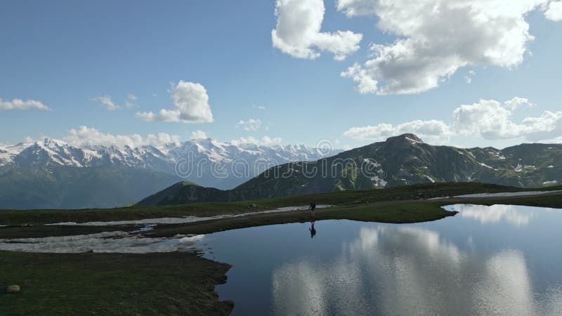 Hiker with backpack walks by Koruldi lakes in Svaneti mountains. Aerial view of snowy peaks, green valleys, clear sky stock video