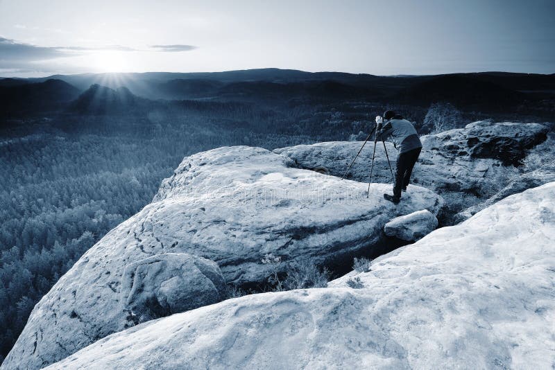 Hiker with camera on tripod takes picture from rocky summit. Alone photographer at edge photograph misty landscape colorful forest in valley, working, wet, weaather, water, trekking, sun, success, stone, spring, sky, sandstone, rocks, rays, rain, professional, peak, park, outdoor, orange, natural, mountain, morning, man, light, job, jacket, humidity, holiday, hilly, heather, green, fresh, fogy, foggy, fairy, dslr, dream, cracks, condition, cold, bush, bowed, block, bent