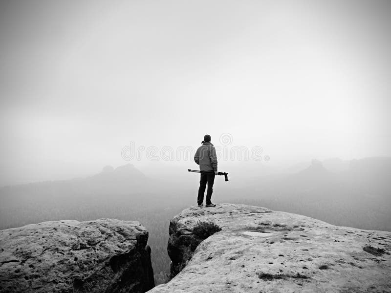 Hiker with camera on tripod takes picture from rocky summit. Alone photographer at edge photograph misty landscape, forest in valley. Hiker with camera on tripod takes picture from rocky summit. Alone photographer at edge photograph misty landscape, forest in valley.