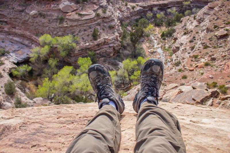 A hiker dangles his feet over the edge of a cliff looking down into a desert canyon in this first person pov photo. A hiker dangles his feet over the edge of a cliff looking down into a desert canyon in this first person pov photo.