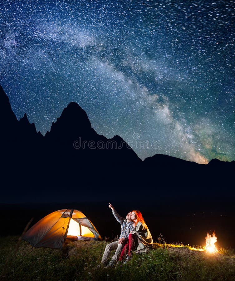 Hiker man showing his lady stars and Milky way in the night sky. Couple sitting near the lighting tent and campfire. royalty free stock photos Hiker man showing his lady stars and Milky way in the night sky. Couple sitting near the lighting tent and campfire. royalty free stock photos