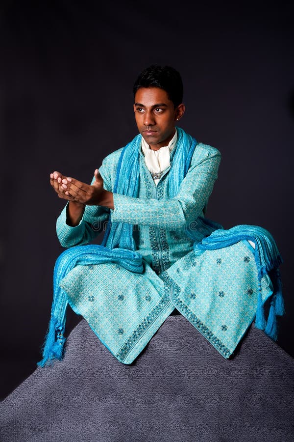 Beautiful authentic Indian hindu man in typical ethnic groom attire sitting in lotus position with legs crossed on top of rock and sticking hands out to receive a gift. Bengali male wearing a light blue agua decorated Dhoti with shawl. Beautiful authentic Indian hindu man in typical ethnic groom attire sitting in lotus position with legs crossed on top of rock and sticking hands out to receive a gift. Bengali male wearing a light blue agua decorated Dhoti with shawl.