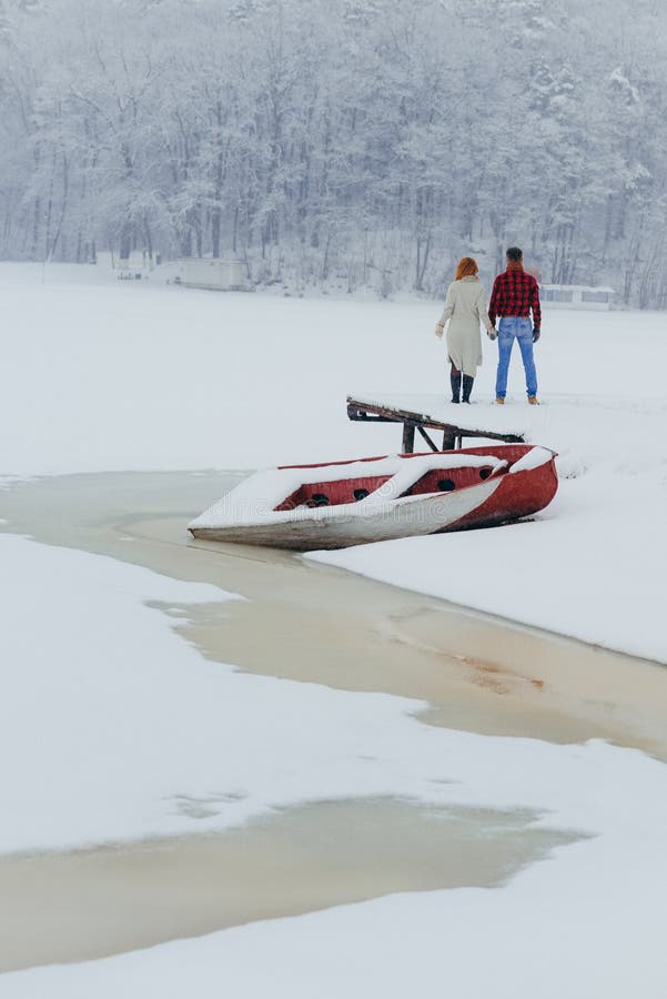 Hintere Ansicht der Vertikale des Paarhändchenhaltens in der Front des Bootes bedeckt mit Schnee im Wald während lizenzfreie stockfotografie Hintere Ansicht der Vertikale des Paarhändchenhaltens in der Front des Bootes bedeckt mit Schnee im Wald während lizenzfreie stockfotografie