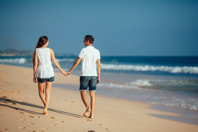Hintere Ansicht des romantischen glücklichen Paars gehend auf Strandhändchenhalten am Hintergrund des blauen Himmels und des Ozea stockfoto Hintere Ansicht des romantischen glücklichen Paars gehend auf Strandhändchenhalten am Hintergrund des blauen Himmels und des Ozea stockfoto