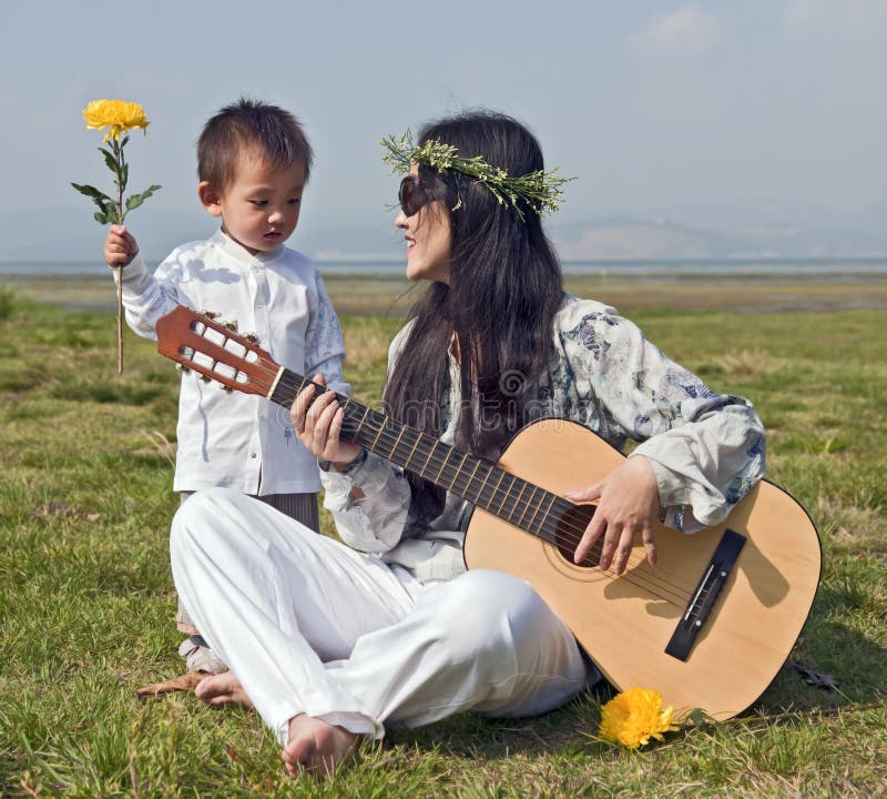 A hippie woman plays the guitar as her son looks on holding a yellow flower. A hippie woman plays the guitar as her son looks on holding a yellow flower.