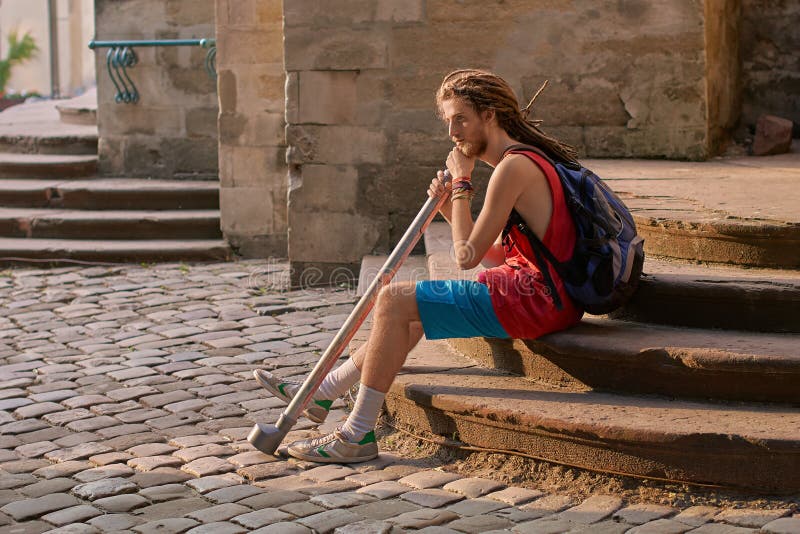 Hippie farpada considerável nova do homem com os dreadlocks que sentam-se com seus trouxa e didgeridoo imagens de stock royalty free Hippie farpada considerável nova do homem com os dreadlocks que sentam-se com seus trouxa e didgeridoo imagens de stock royalty free