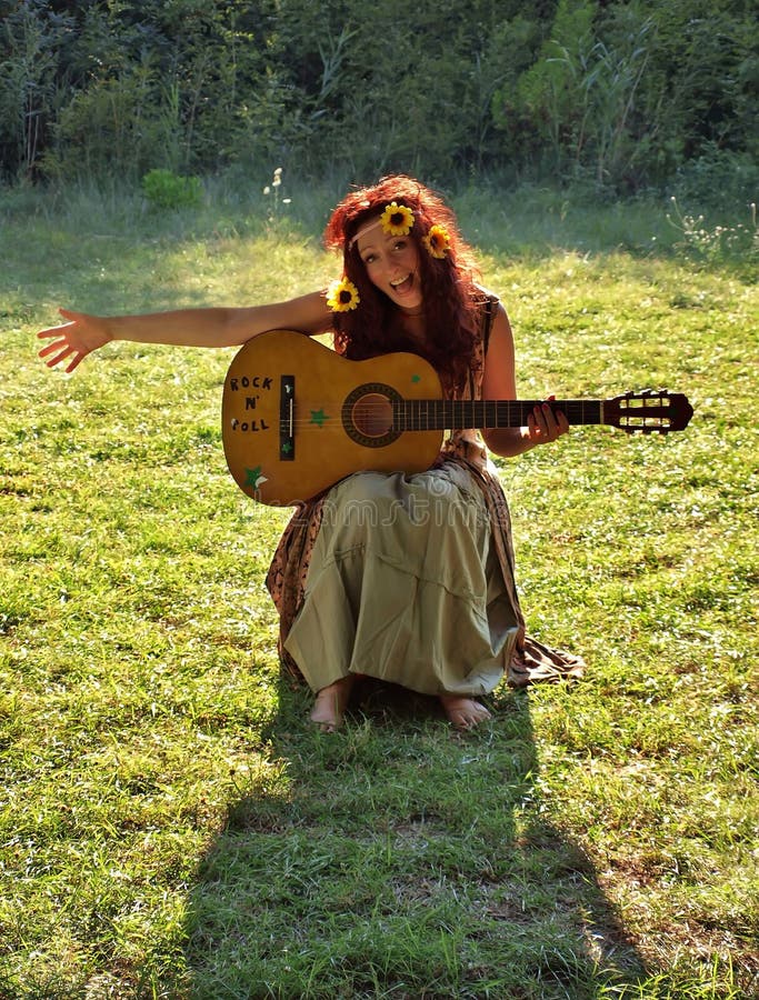 Hezký usměvavá brunetka žena s květy ve vlasech sedí na zelené trávě, hrát na kytaru a zpívá píseň.