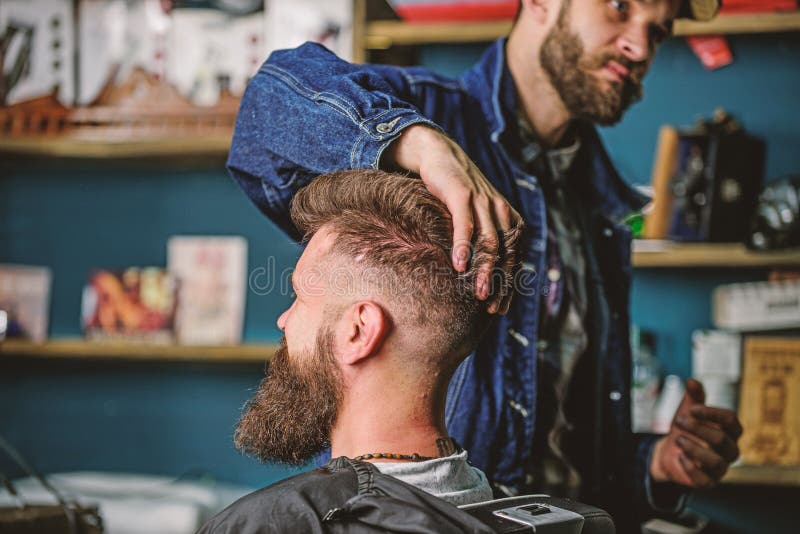 Hipster client with fresh haircut or hairstyle. Barber styling hair of bearded client with wax by hands. Man with beard