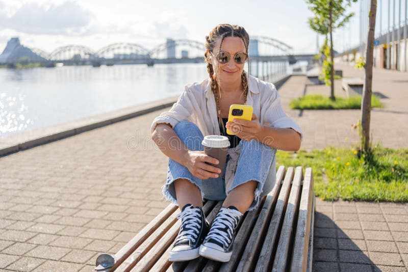 hipster female with braids and takeaway coffee sitting using smartphone at street of city. Happy student girl sitting outside with smartphone in hands having coffee break have video call with friends. hipster female with braids and takeaway coffee sitting using smartphone at street of city. Happy student girl sitting outside with smartphone in hands having coffee break have video call with friends.