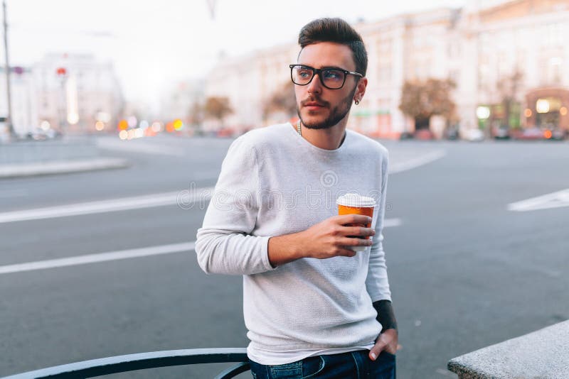 Hipster man standing with takeaway coffee, smiling plesantly, walking on th city street. Happy carefree handsome guy in eyewear