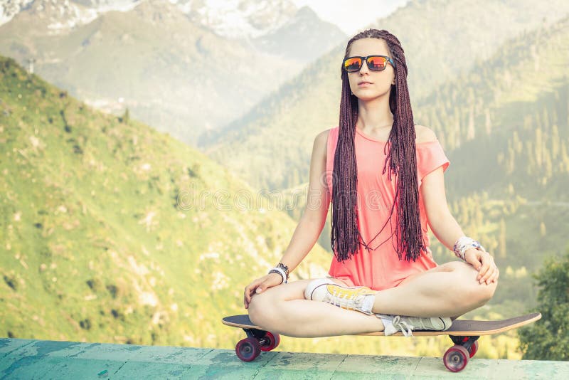 Hipstermodeflicka som gör yoga som kopplar av på skateboarden på berget royaltyfria foton Hipstermodeflicka som gör yoga som kopplar av på skateboarden på berget royaltyfria foton
