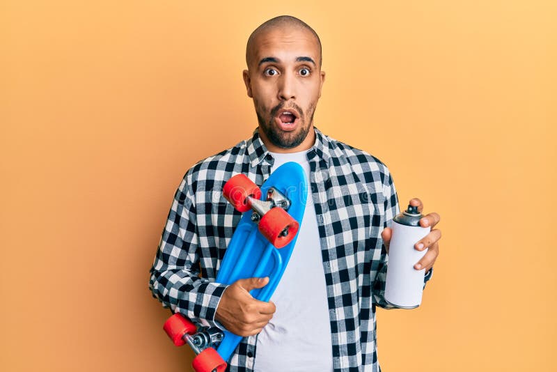 Hispanic adult skater man holding skate and graffiti spray afraid and shocked with surprise and amazed expression, fear and excited face